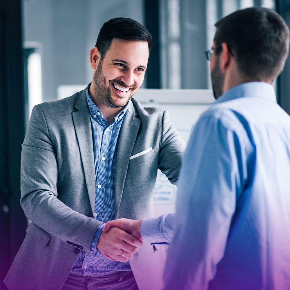 Dois homens brancos apertando as mãos. Um deles usa um terno cinza e camisa azul, o outro uma camisa azul. Eles estão em um ambiente empresarial e o gesto representa o negócio fechado.