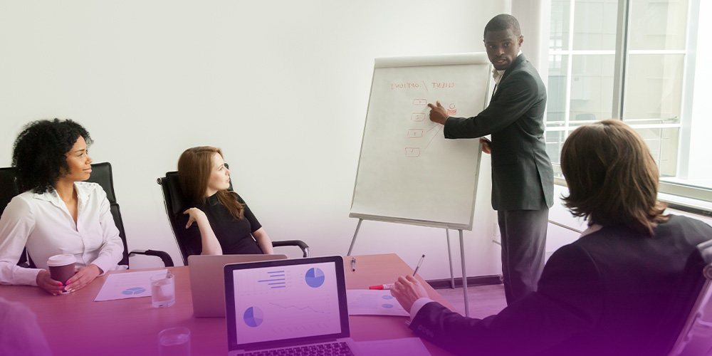 A imagem representa a equipe de marketing definindo estratégias. Nela, um homem negro, usando um terno preto, aponta para um quadro com gráficos. Duas mulheres e um homem sentados em uma mesa, um homem e duas mulheres, olham para ele. Tem um notebook com gráficos na tela ao lado de uma das pessoas.