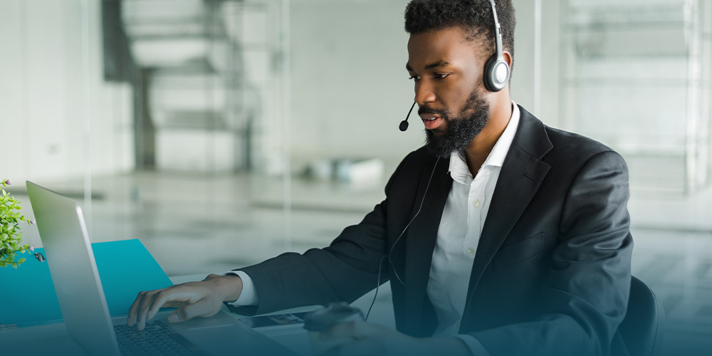 Um homem negro, usando camisa branca e paletó preto, está usando um headset e em frente a um notebook, representando a prospecção ativa, ou seja, o outbound marketing.